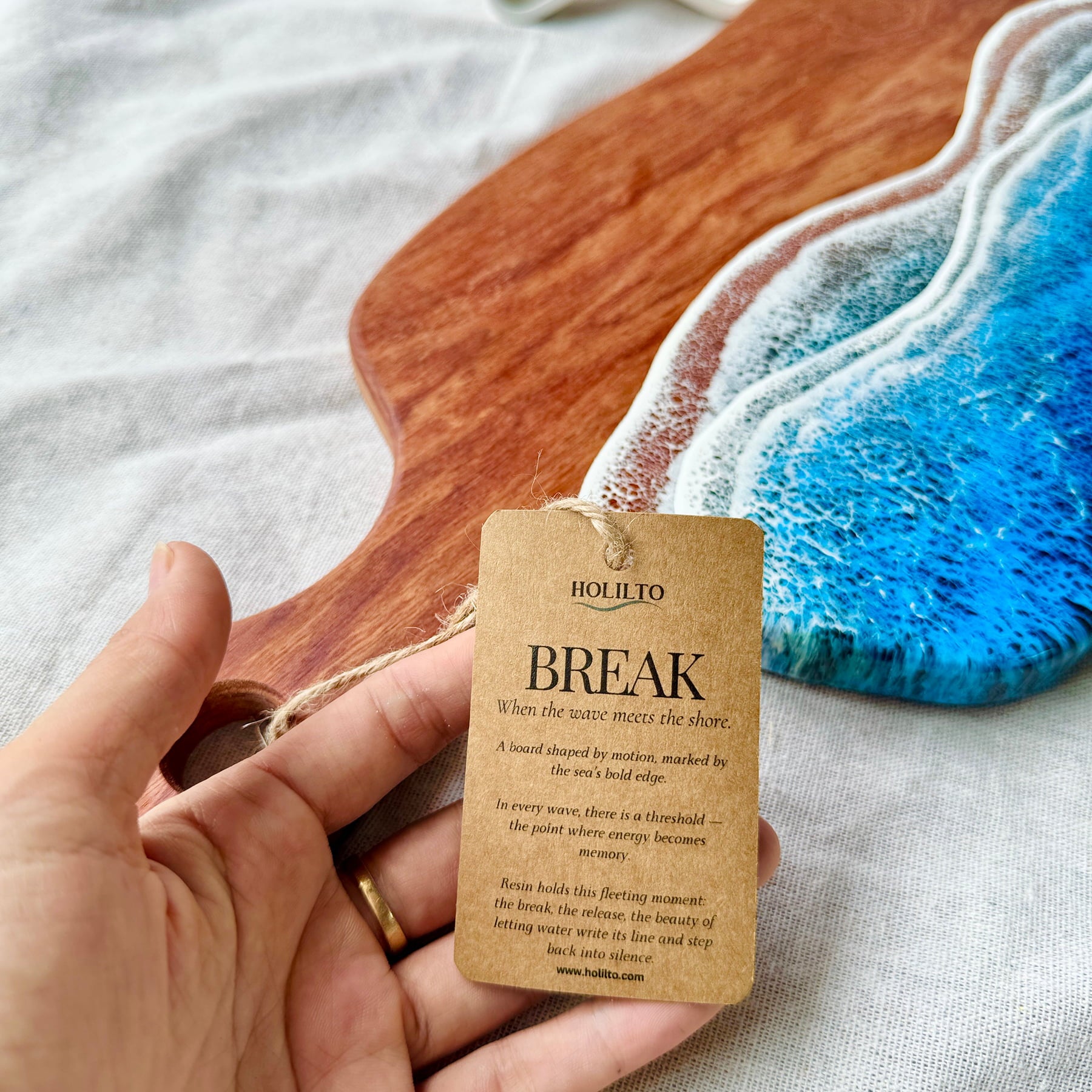 Hand holding a 'Holilto' tag with a wooden and resin artwork in the background