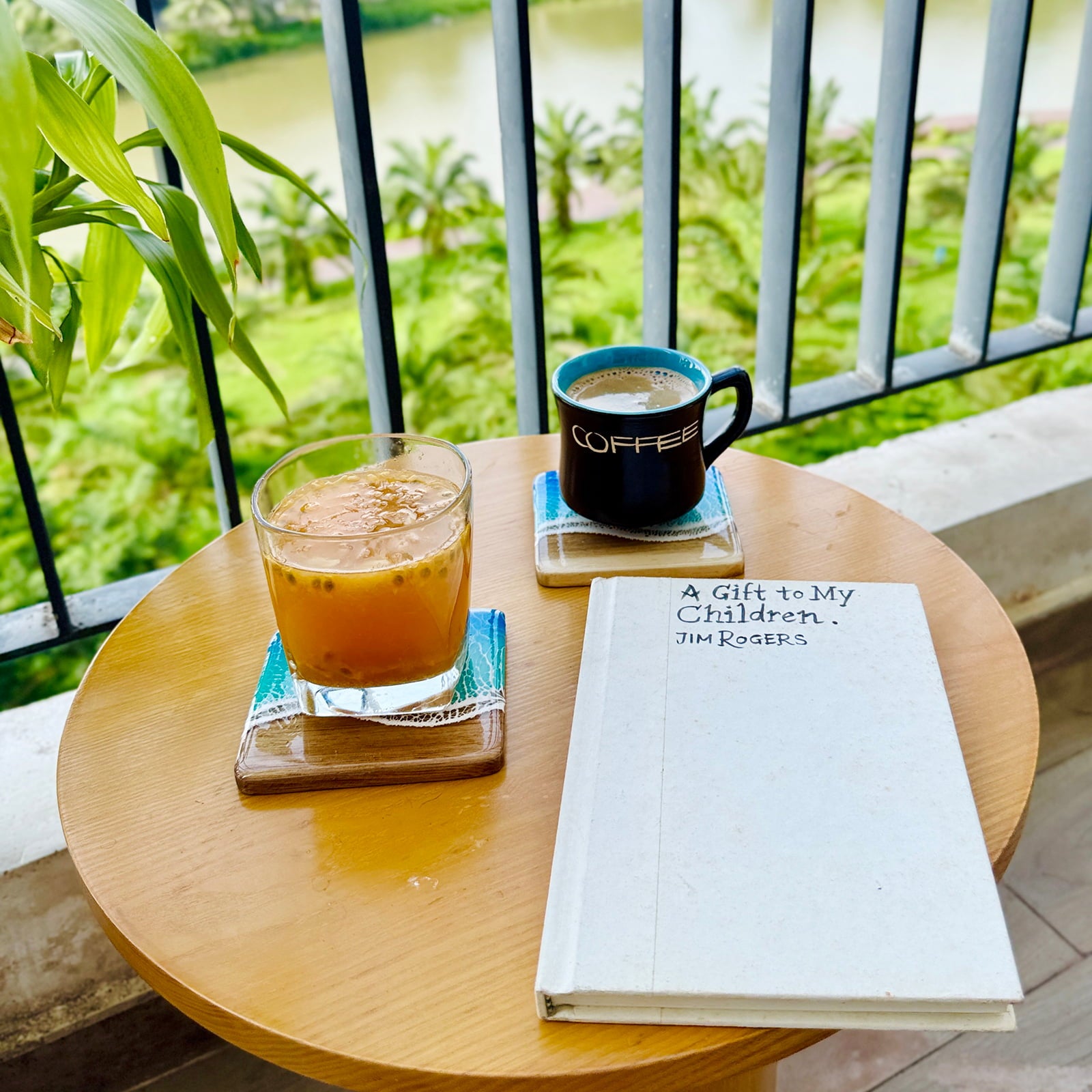 Table with a book, coffee cup, and glass of iced tea on a balcony with greenery.