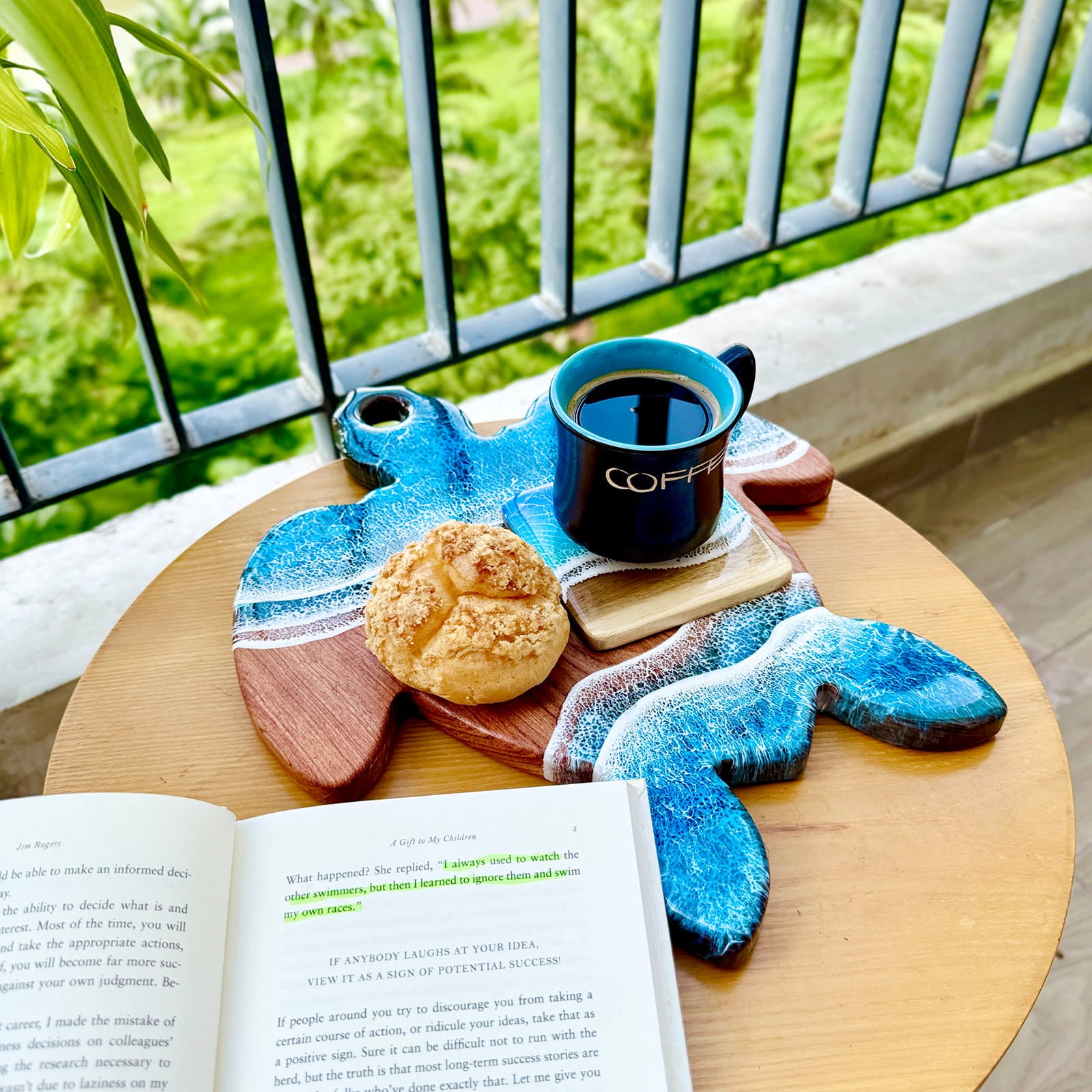 Tray with a turtle design holding a cup of coffee, pastry, and book on a wooden table outdoors.